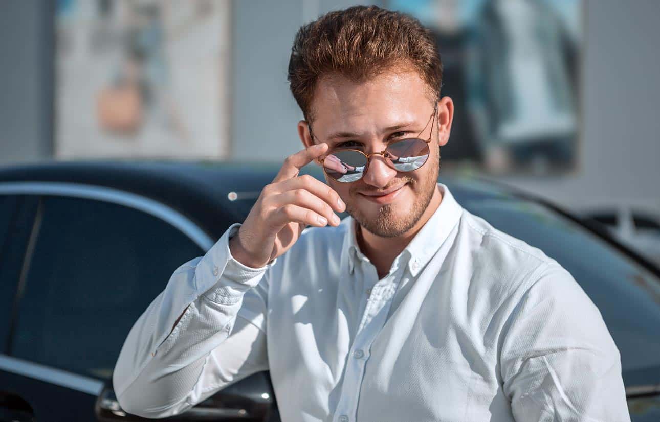 hipster-salesman-with-aviator-glasses-standing-in-front-of-luxury-car-signifying-deceit-and-greed