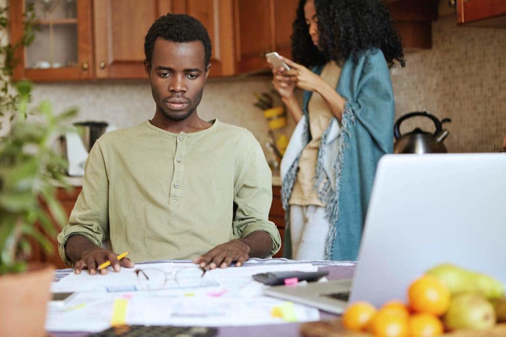 husband-sitting-at-table-looking-at-unbearable-bills-wondering-what-to-do-with-timeshare-wife-in-kitchen-on-smartphone