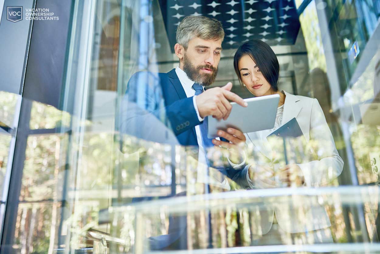 middle-aged-man-in-suit-explaining-process-on-tablet-to-younger-woman-looking-out-window-where-photograher-is-standing-looking-up