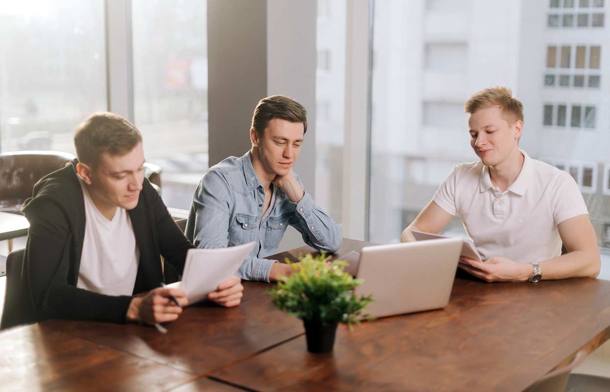 young-men-sitting-around-conference-room-table-doing-research-on-computer-scheming-for-sales-presentation-for-vacations