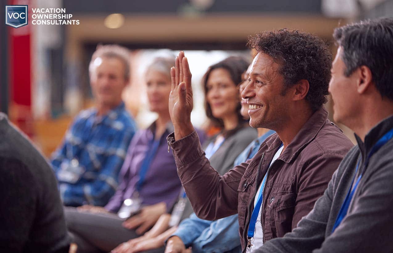 middle-aged-black-man-raising-hand-at-timeshare-seminar-demonstration-with-smirk-on-face-as-if-he's-got-a-good-question-to-ask-in-front-of-group